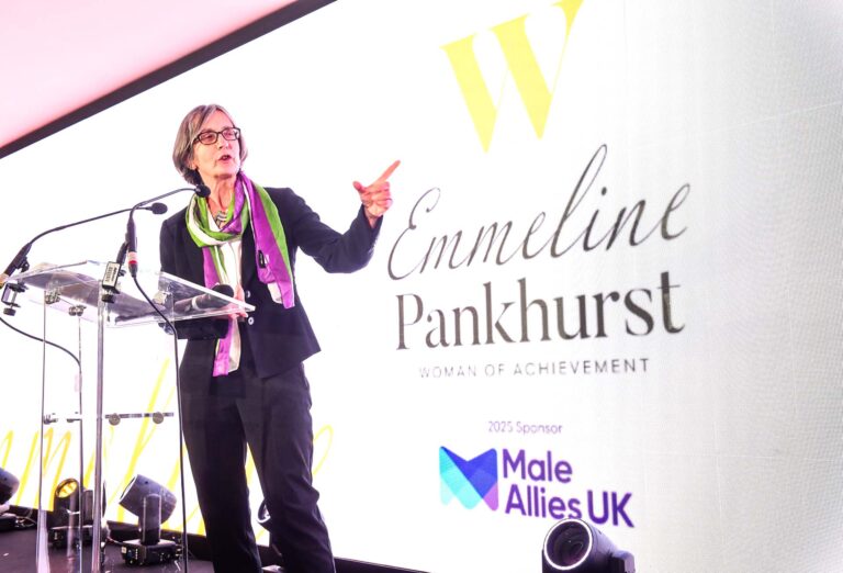 A woman wearing a scarf of the suffragette colours - white, green and purple - standing in front of a screen that reads 'Emmeline Pankhurst woman of achievement 2025 sponsor Male Allies UK'