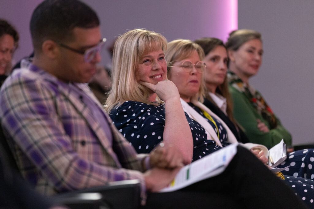 A close up of a blonde woman smiling while listening to the speakers on stage, looking really engaged. From the National Allyship Summit