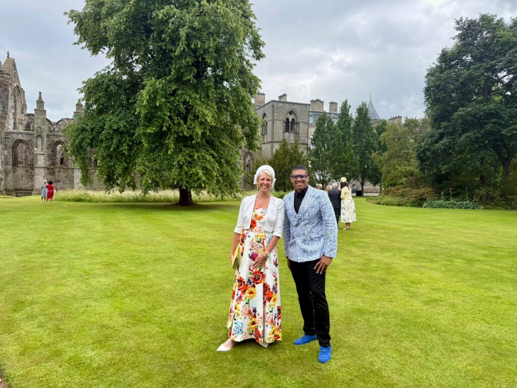 Lee Chambers is wearing a light blue floral blazer and blue shoes smiling standing next to Anna Price in a white and red floral dress and bolero. The Palace of Holyrood house and lush gardens is behind them.